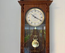 Acctim quartz battery-operated pendulum regulator wall clock housed in a carved wooden case with a dark oak finish.