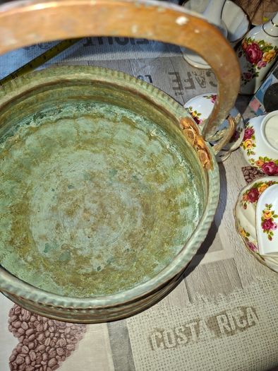 Antique Copper Cauldron features a Rustic Patina, a Swing Handle, and Distinctive Ribbed Detailing around the Rim. It resembles vintage metal pots often used for hearth decoration or as Planters. The piece shows significant oxidation and aging, characteri