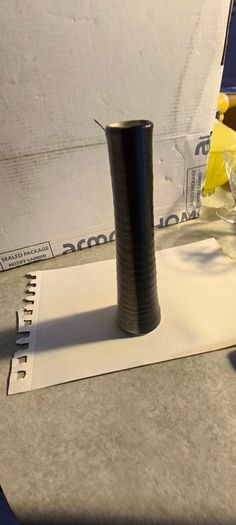 Tall, slender, textured cylindrical ceramic or wooden decorative object with a two-toned vertical or striped design, resembling modern art pottery or a candle holder.