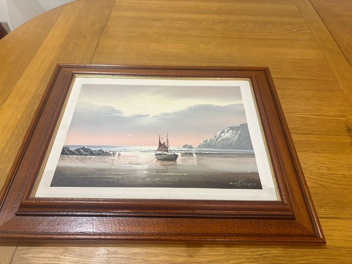 This artwork is a coastal oil painting featuring a traditional single-masted sailing boat stranded or anchored on a calm shoreline, signed by contemporary marine artist David James