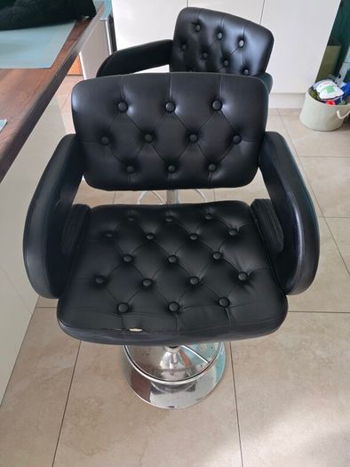 2 black faux leather bar stools with padded seats, armrests and chrome bases.
