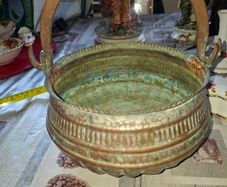 Antique Copper Cauldron features a Rustic Patina, a Swing Handle, and Distinctive Ribbed Detailing around the Rim. It resembles vintage metal pots often used for hearth decoration or as Planters. The piece shows significant oxidation and aging, characteri