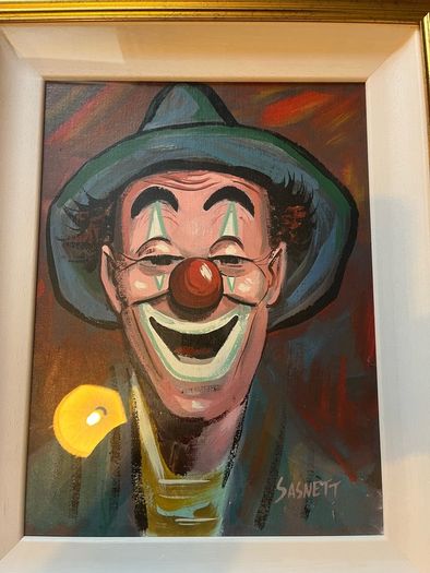 Portrait of a smiling clown wearing a green brimmed hat, red nose, and blue/green face paint around the eyes and mouth.