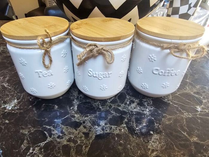 white ceramic tea, coffee, and sugar storage canisters accented with embossed daisy patterns and rustic twine bows.