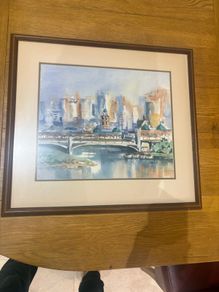 The framed watercolor painting appears to depict an impressionistic cityscape featuring a historic stone bridge over a river, similar in style and subject to watercolor prints of Princes Bridge in Melbourne or the Melbourne Skyline with Yarra River