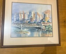 The framed watercolor painting appears to depict an impressionistic cityscape featuring a historic stone bridge over a river, similar in style and subject to watercolor prints of Princes Bridge in Melbourne or the Melbourne Skyline with Yarra River
