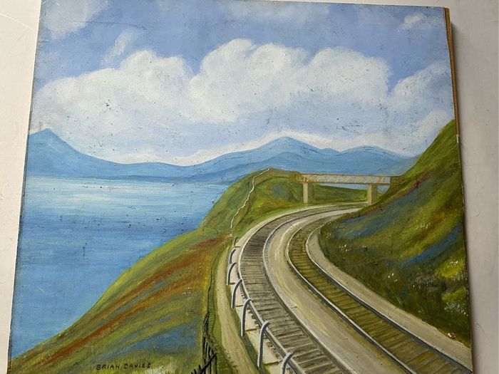 picture This piece depicts a railway line sweeping around a coastal bay beneath a footbridge and hillsides, signed Brian Davies in the lower left corner.