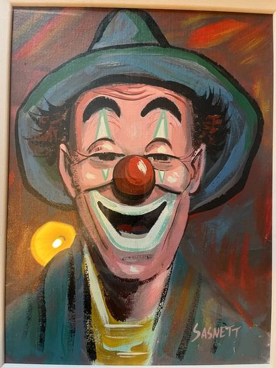 Portrait of a smiling clown wearing a green brimmed hat, red nose, and blue/green face paint around the eyes and mouth.