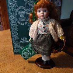 Collectors porcelain Irish doll from the "Collectors Dolls" range by Shannon Distributors. It is specifically marketed as a doll featuring traditional Irish dresses, as indicated by the tag it's holding. It is new in box.