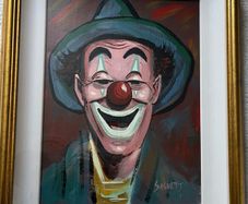 Portrait of a smiling clown wearing a green brimmed hat, red nose, and blue/green face paint around the eyes and mouth.