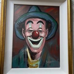 Portrait of a smiling clown wearing a green brimmed hat, red nose, and blue/green face paint around the eyes and mouth.