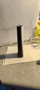 Tall, slender, textured cylindrical ceramic or wooden decorative object with a two-toned vertical or striped design, resembling modern art pottery or a candle holder.