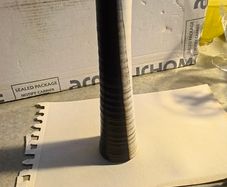 Tall, slender, textured cylindrical ceramic or wooden decorative object with a two-toned vertical or striped design, resembling modern art pottery or a candle holder.