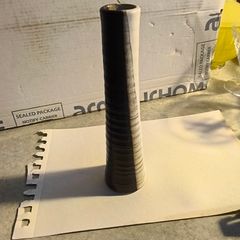 Tall, slender, textured cylindrical ceramic or wooden decorative object with a two-toned vertical or striped design, resembling modern art pottery or a candle holder.