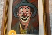 Portrait of a smiling clown wearing a green brimmed hat, red nose, and blue/green face paint around the eyes and mouth.