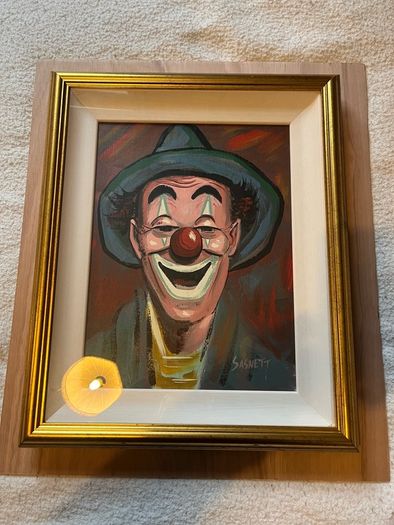 Portrait of a smiling clown wearing a green brimmed hat, red nose, and blue/green face paint around the eyes and mouth.