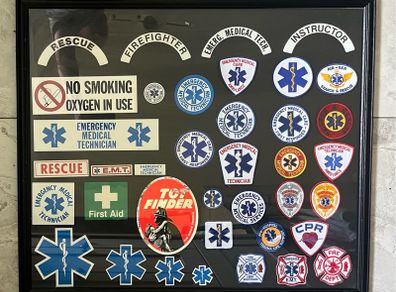Framed display of EMS (Emergency Medical Services) including Fire and Ambulance Service Patches and stickers. Frame measures 34 incehs X 29 inches
