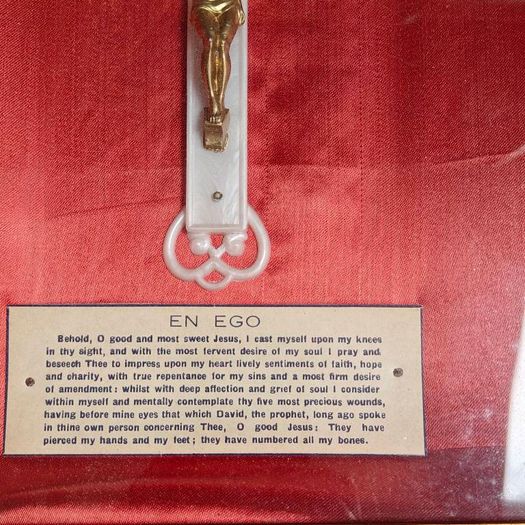antique framed crucifix featuring a white ornamental cross with a metallic corpus of Jesus Christ mounted over a red  backing inside a gilded wooden frame. Devotional item.