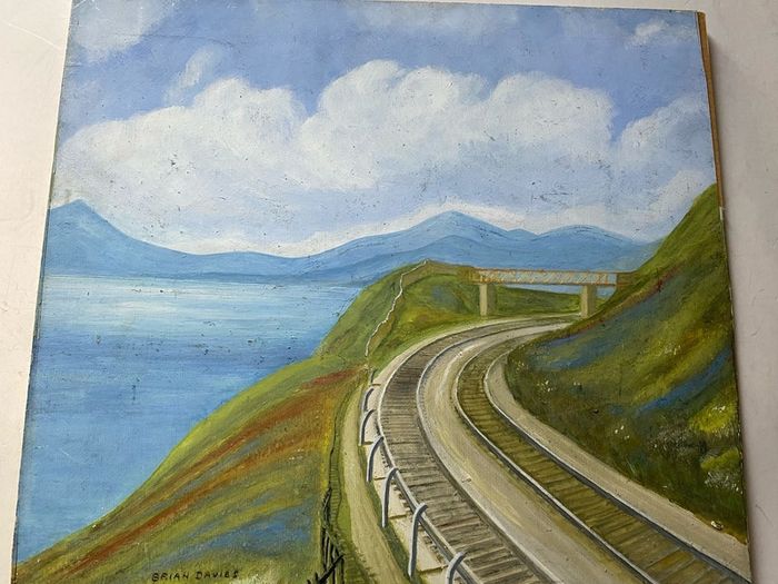 picture This piece depicts a railway line sweeping around a coastal bay beneath a footbridge and hillsides, signed Brian Davies in the lower left corner.